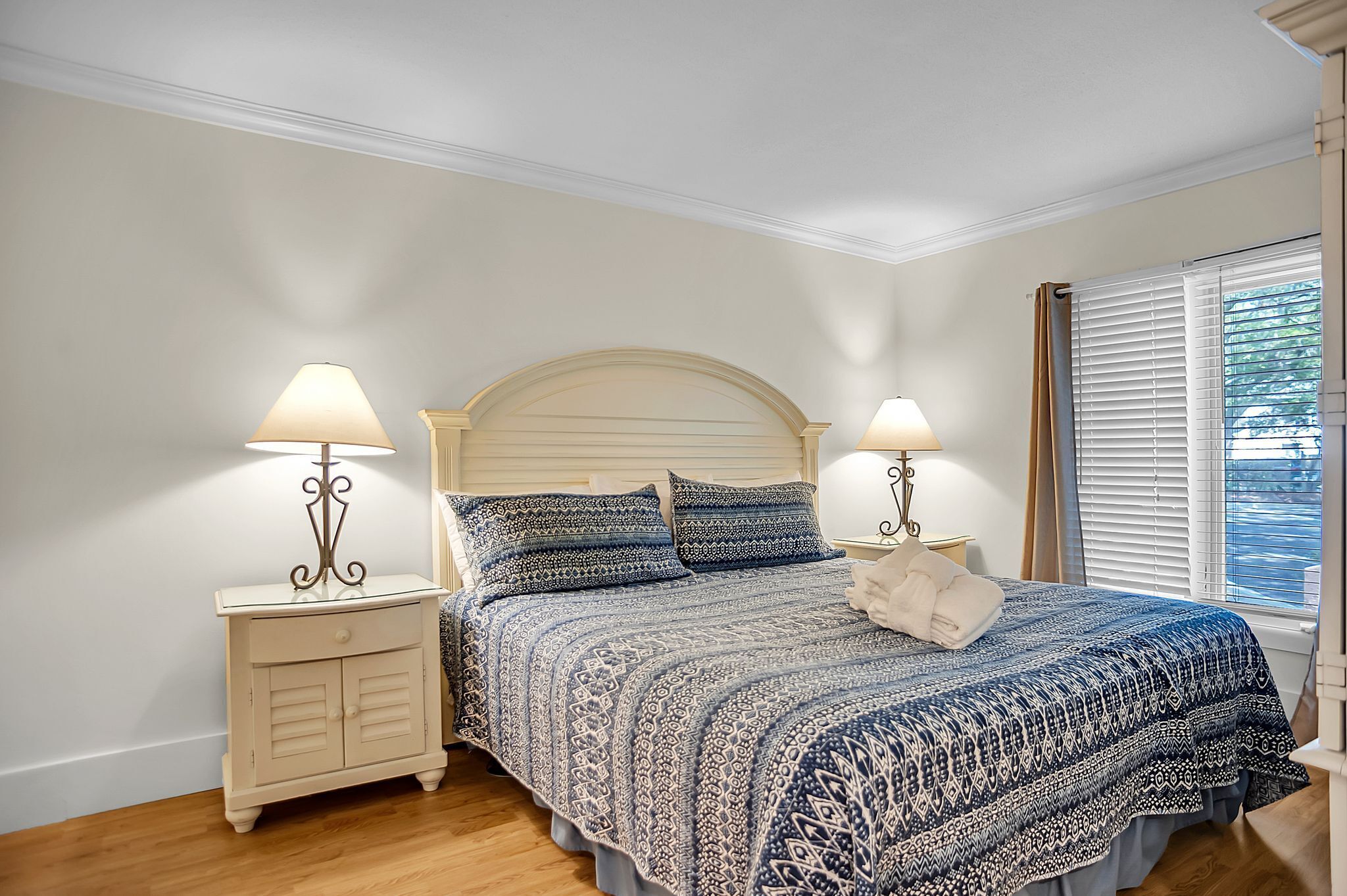A bedroom with a bed, two bedside tables with lamps, a window with blinds, and a wooden floor.