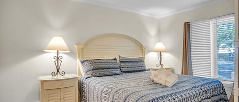 A bedroom with a bed, two bedside tables with lamps, a window with blinds, and a wooden floor.