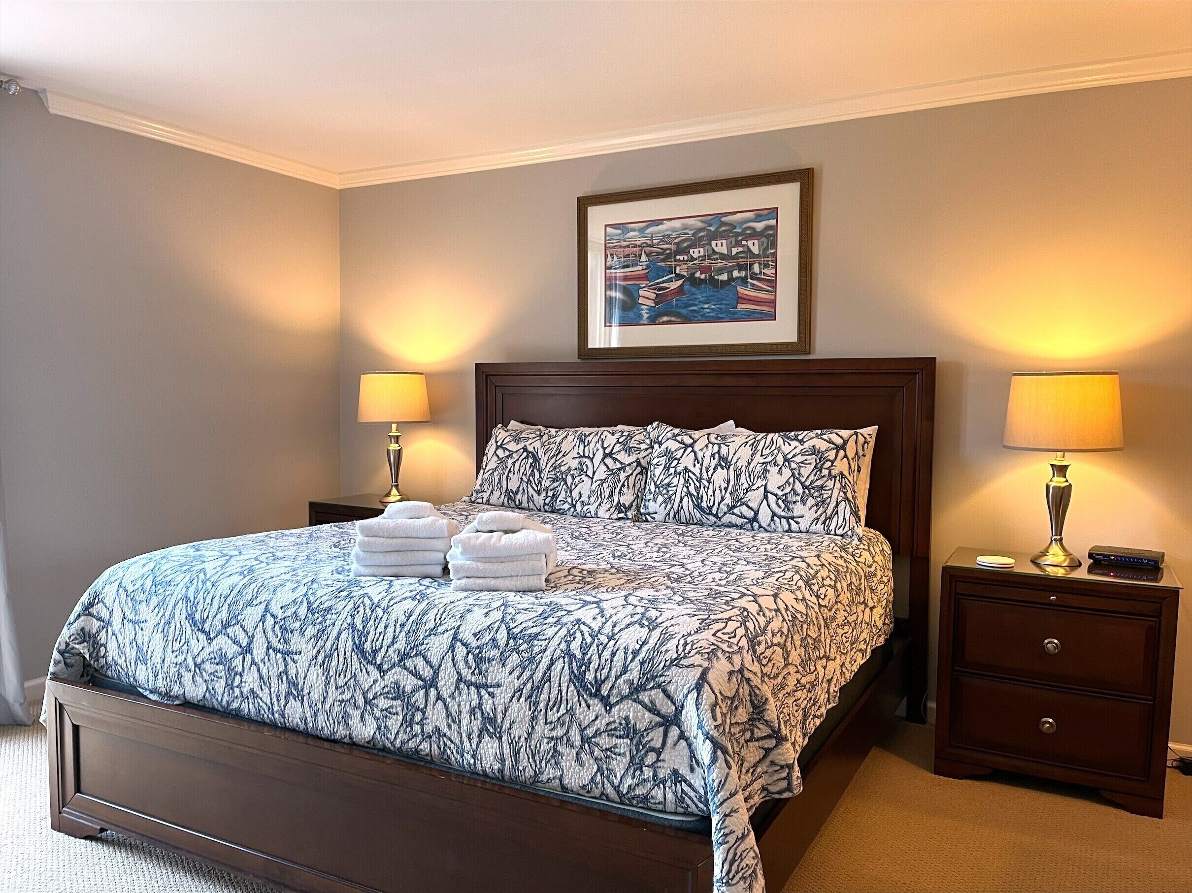 A neatly made bed with a patterned bedspread, two bedside lamps, a framed picture above the bed, and a nightstand with a remote control.