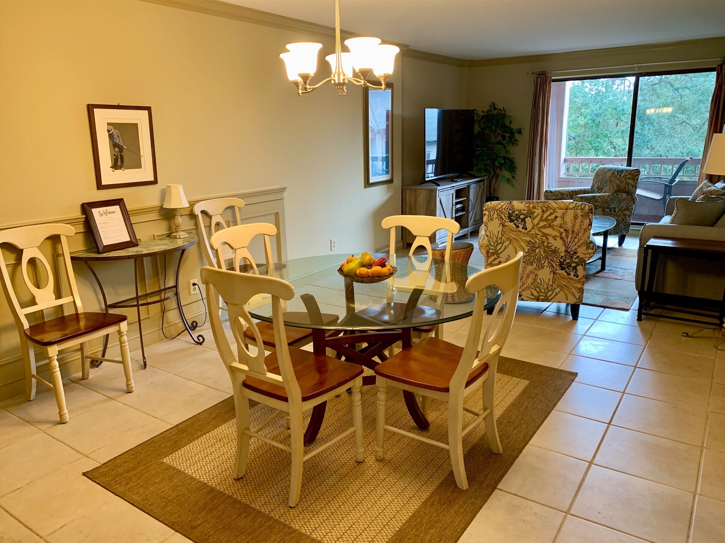 A dining area with a glass table, white chairs, and a fruit bowl. There is a TV and a plant in the background.