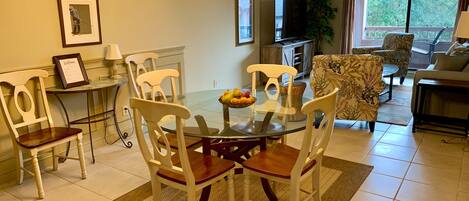 A dining area with a glass table, white chairs, and a fruit bowl. There is a TV and a plant in the background.