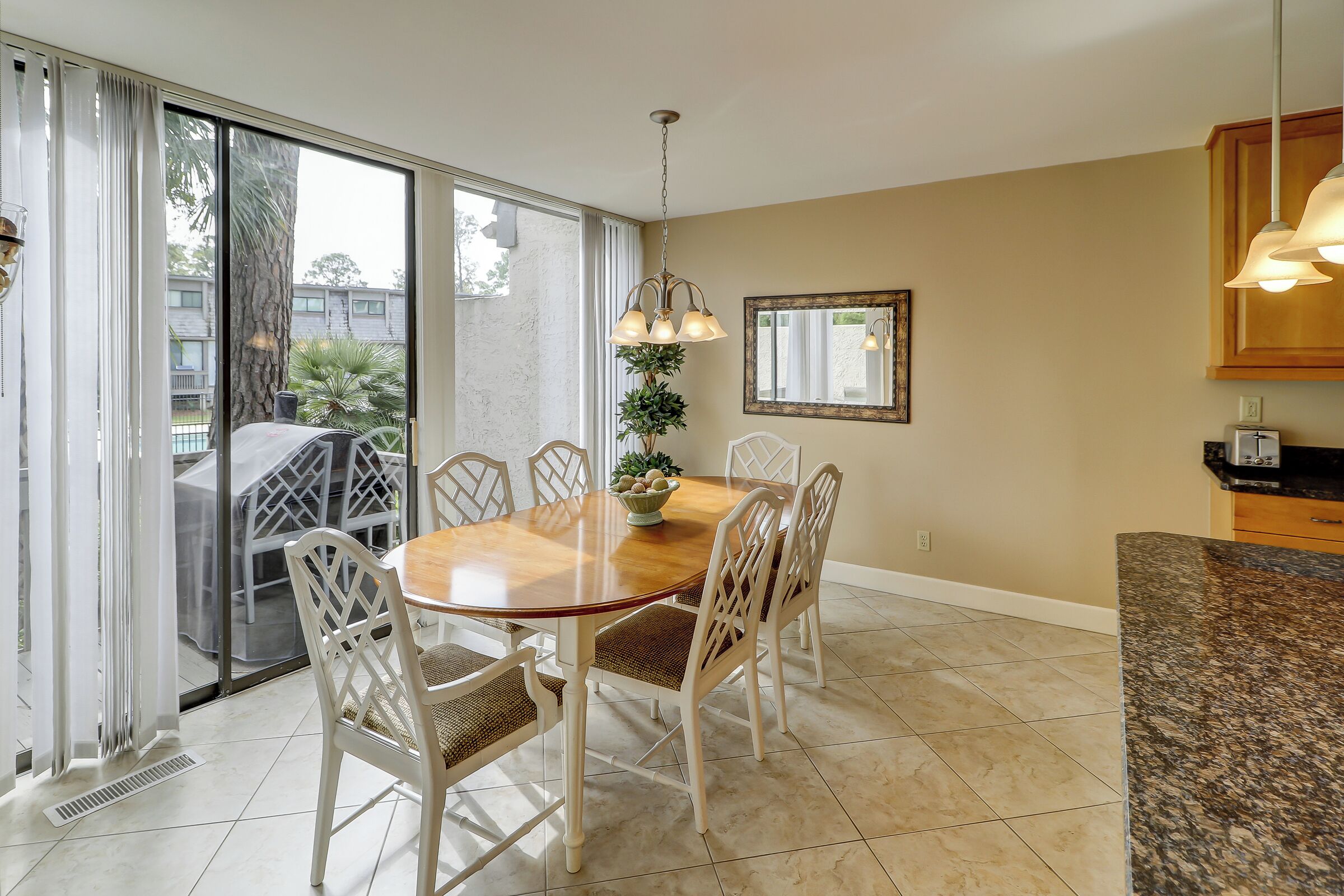 A dining area with a wooden table and white chairs, a ceiling light, and a view through large glass doors.