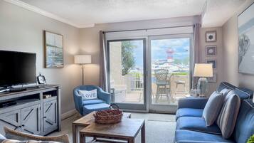 A living room with a TV, a blue armchair with a LOVE pillow, a blue sofa, a wooden coffee table, and a view of boats and a lighthouse through the glass door.