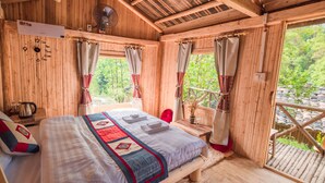 Una habitación tipo cabaña de madera con una cama, un ventilador, una ventana con cortinas y vistas al follaje exterior.
