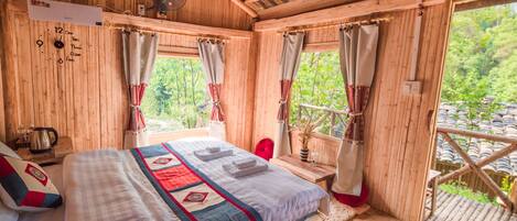 A wooden cabin room with a bed, a fan, a window with curtains, and a view of greenery outside.