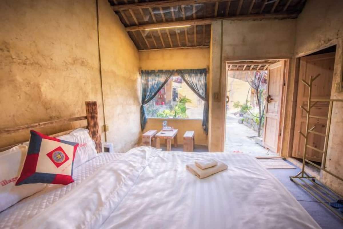 A room with a bed, a window with curtains, a small table, and a wooden door.