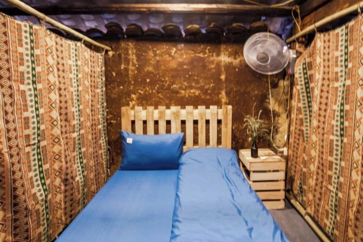 A small, cozy room with a wooden headboard, a single bed with blue bedding, and patterned curtains.