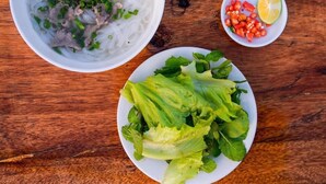 Un tazón de sopa de fideos con carne y cebollas verdes, un plato de lechuga fresca y un recipiente pequeño con tomates en rodajas y lima.
