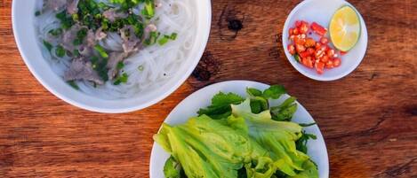 A bowl of noodle soup with meat and green onions, a plate of fresh lettuce, and a small bowl of sliced tomatoes and lime.