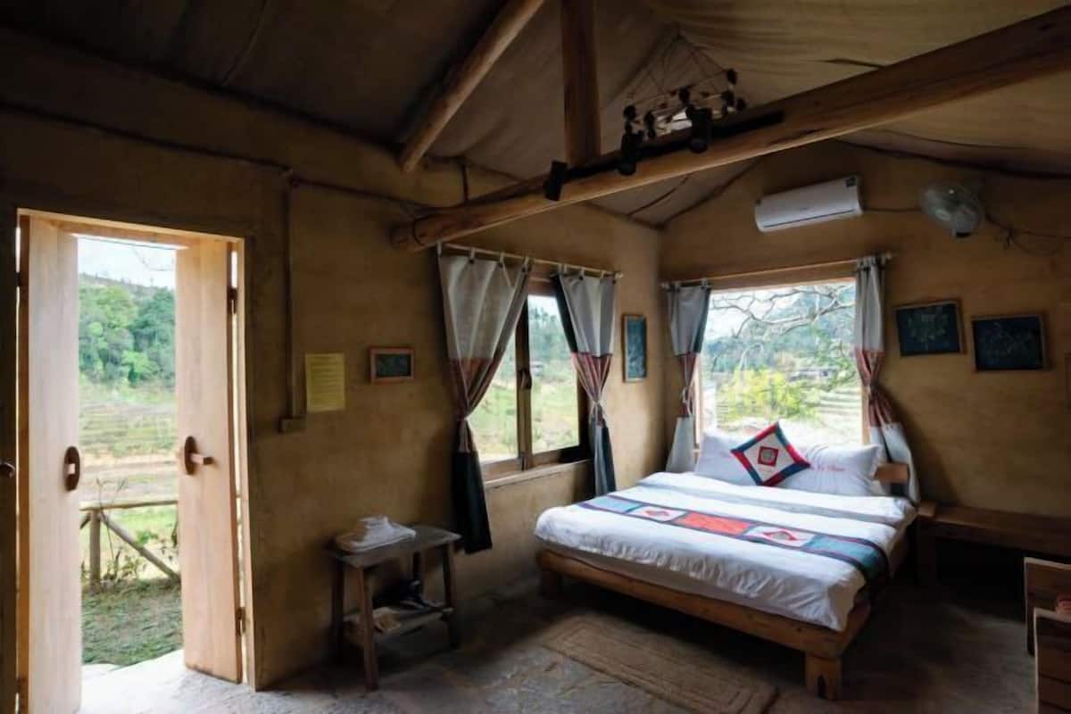 A bedroom with a wooden bed, a small table, a window with curtains, and an open door leading outside.
