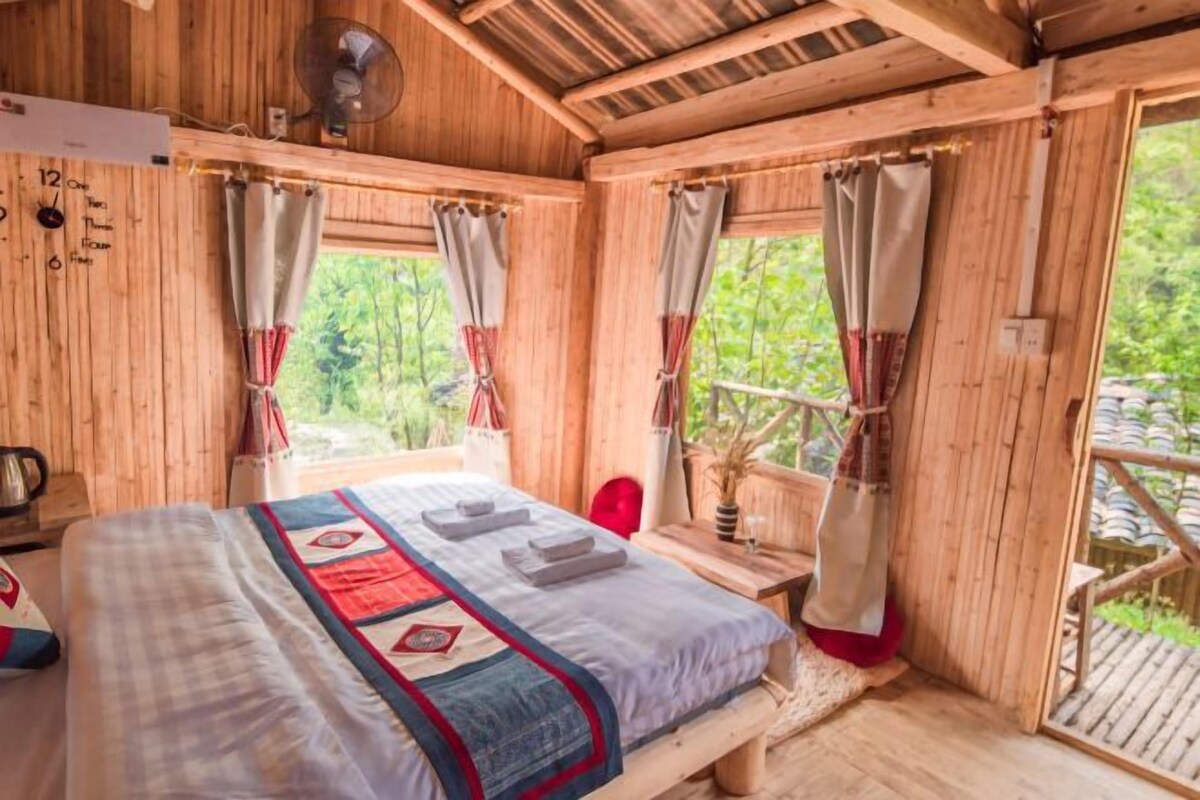 A wooden cabin room with a bed, curtains, a fan, and a window overlooking greenery.