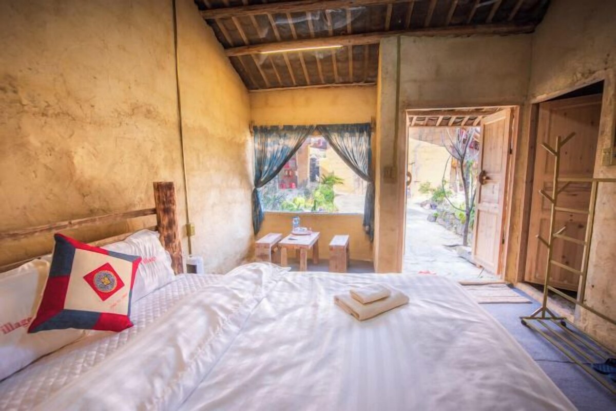 A room with a bed, a window with curtains, a small table, and a wooden door.