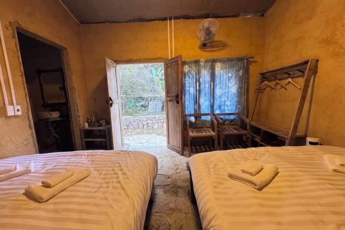 A room with two beds, a fan, a wooden chair, a small table, and a view of the outdoors.