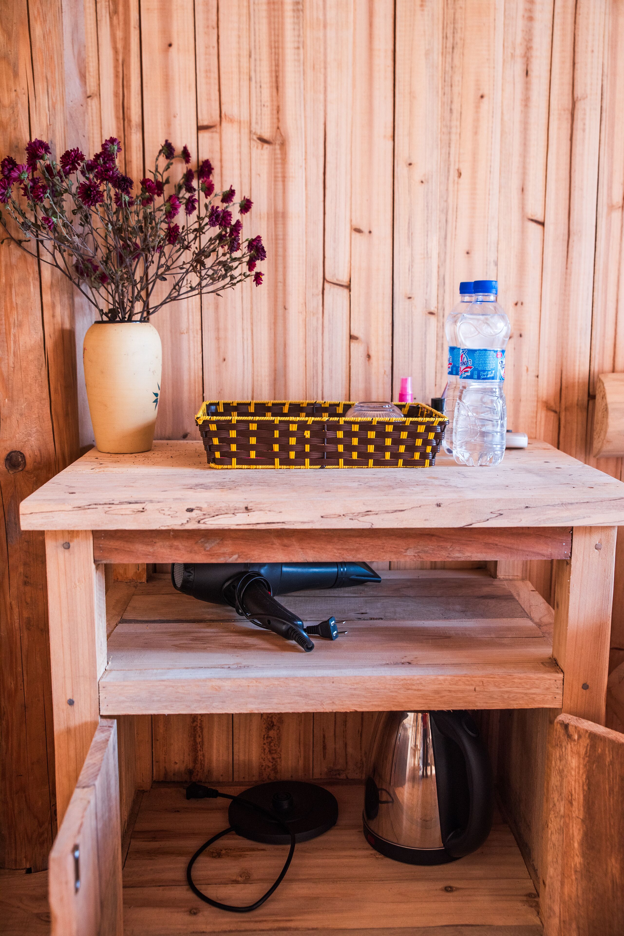 A wooden table with a vase of flowers, a woven basket, a water bottle, and a kettle.
