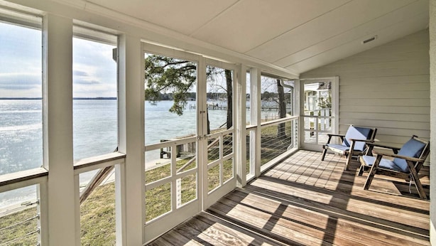 A covered porch with wooden flooring and white railings, overlooking a body of water with a dock and trees.