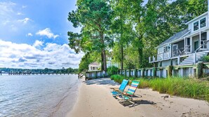 A beach with a wooden pier extending into the water, a house with a porch, and a row of beach chairs along the shoreline.
