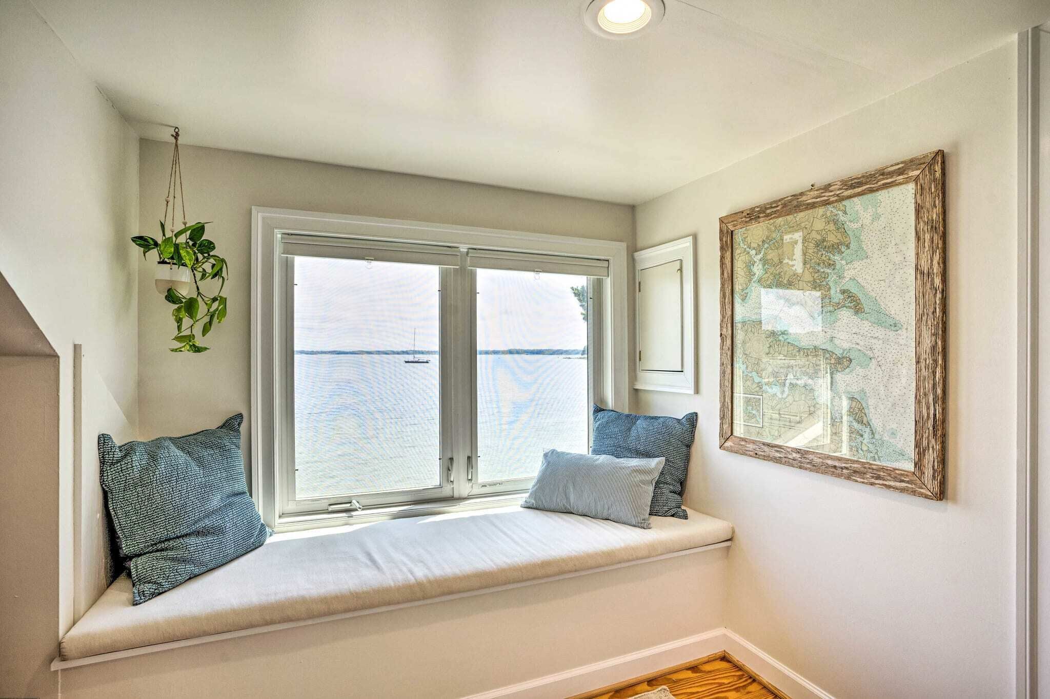 Ein Zimmer mit einer eingebauten Sitzbank an einem großen Fenster mit Blick auf ein Gewässer, einer hängenden Pflanze und einer gerahmten Karte an der Wand.