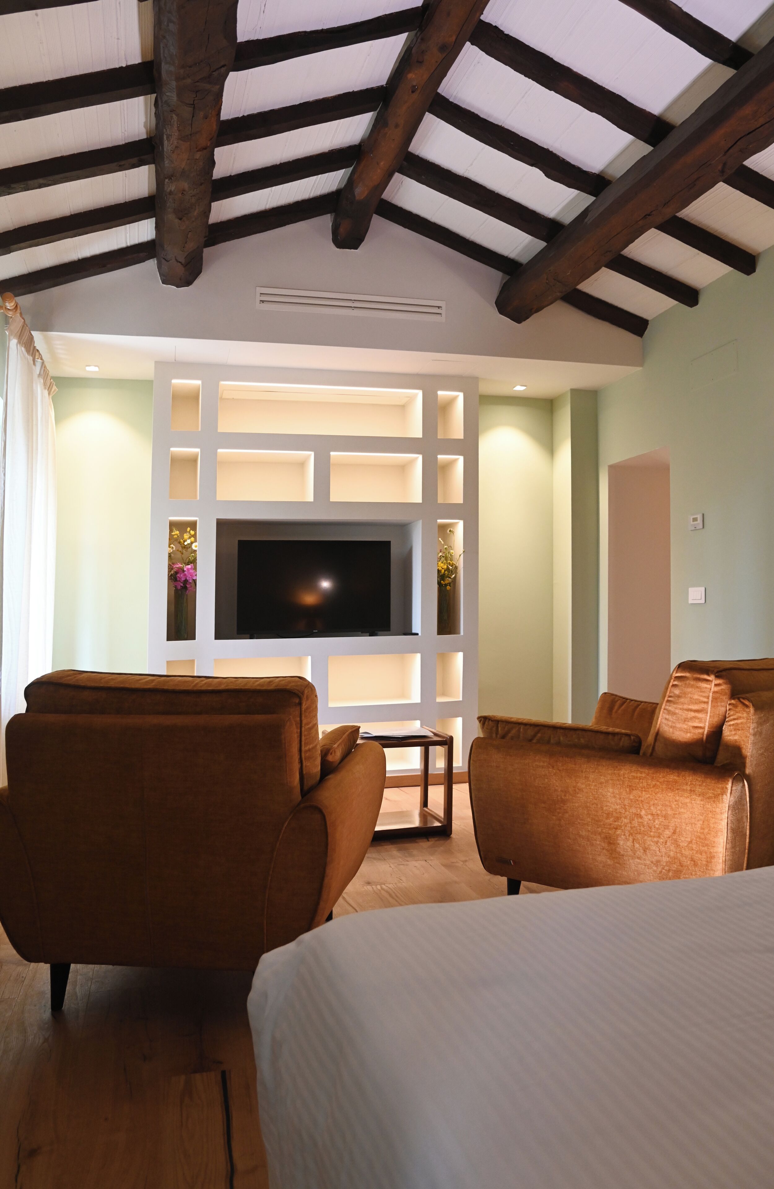 A hotel room with a large flat-screen TV, two brown leather sofas, a bed with white bedding, and a wooden ceiling with exposed beams.