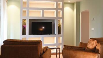 A hotel room with a large flat-screen TV, two brown leather sofas, a bed with white bedding, and a wooden ceiling with exposed beams.
