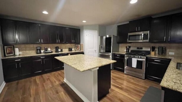 A modern kitchen with dark cabinets, granite countertops, stainless steel appliances, and a wooden floor. - Play & Relax — 4 Kings, Pool Table & 5 TVs Near JW Marriott TPC (San Antonio)