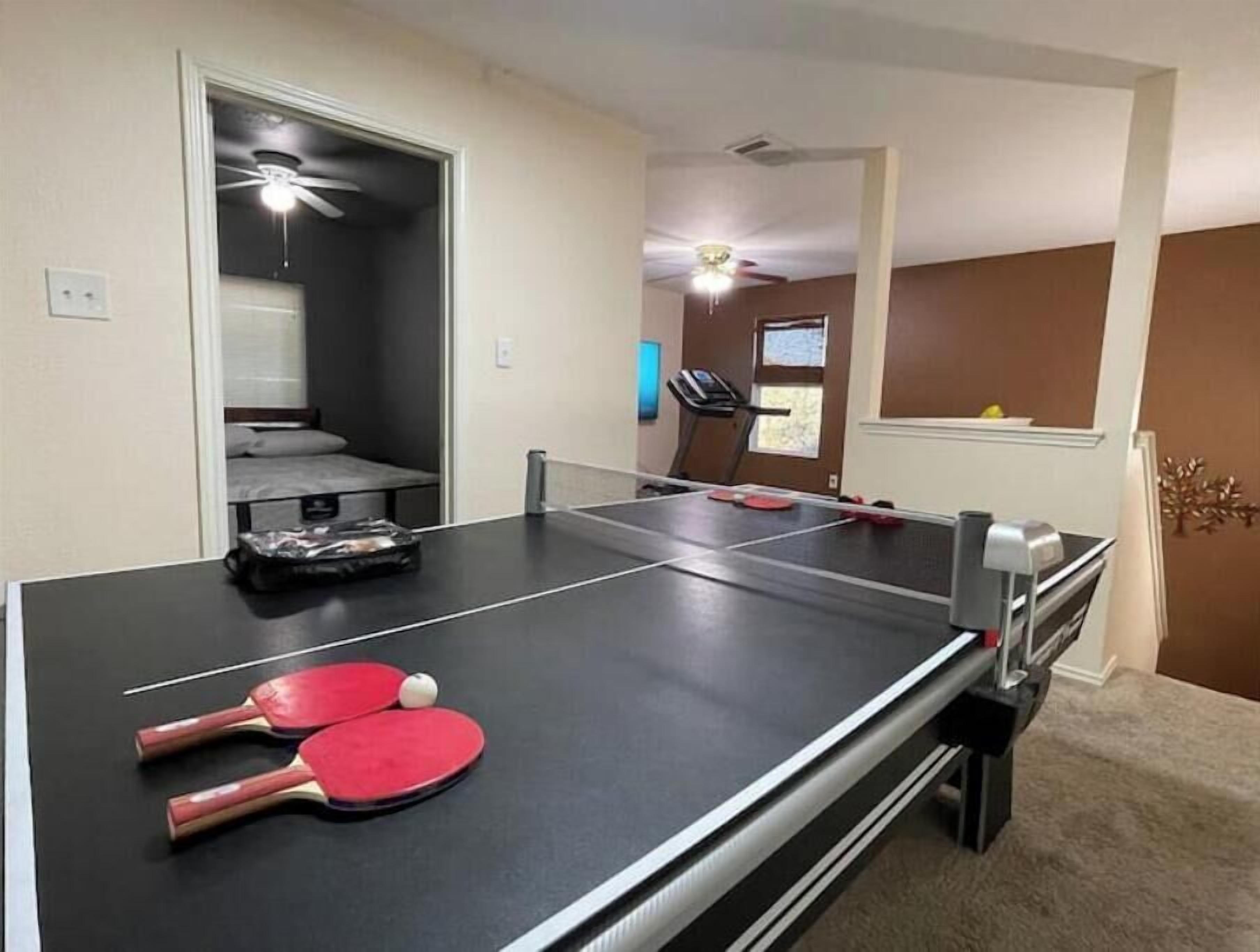 A ping pong table with two red paddles and a ball, a treadmill, and a ceiling fan in a room with beige walls and a carpeted floor.