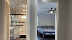 A modern bathroom with a glass shower enclosure and a separate toilet. A bedroom with a bed and a ceiling fan is visible through an open door.