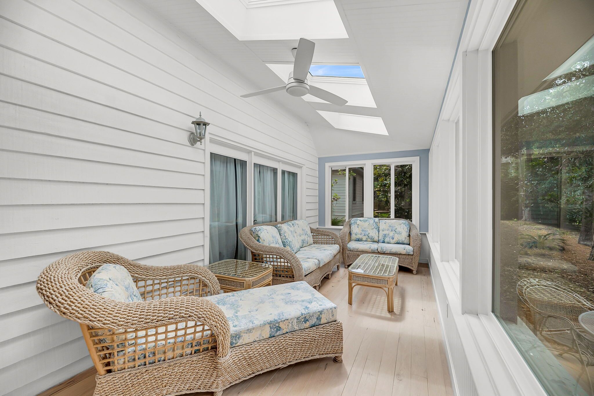 Un patio soleado con mobiliario de mimbre, un ventilador de techo y amplios ventanales con vistas al exterior.