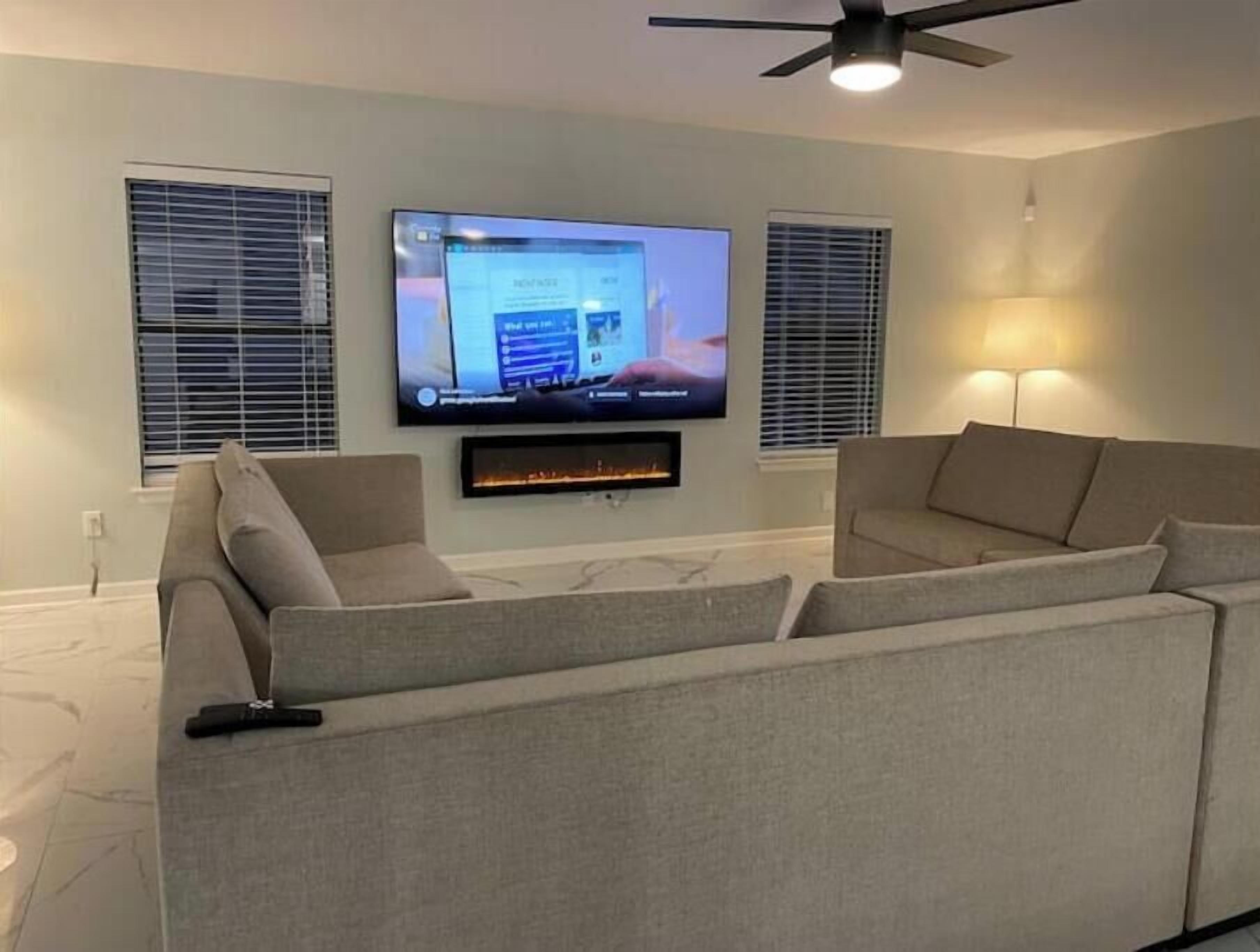 Una sala de estar moderna con un televisor de pantalla plana montado en la pared, una chimenea debajo y dos ventanas con persianas.