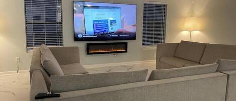 Una sala de estar moderna con un televisor de pantalla plana montado en la pared, una chimenea debajo y dos ventanas con persianas.