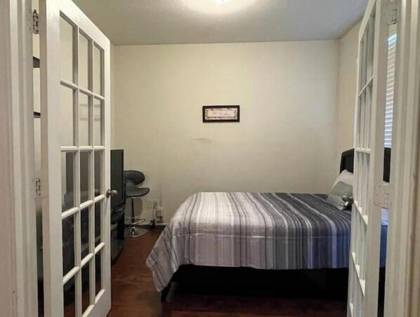 Una habitación pequeña y estrecha con una cama, un escritorio y una silla. Hay un cuadro en la pared y una puerta con paneles de vidrio.