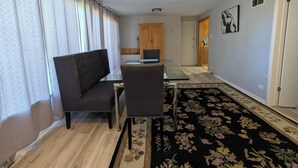 Un área de comedor con una mesa de vidrio, dos sillas tapizadas, un alfombra floral negro y una puerta de madera al fondo.