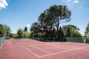 Un court de tennis en plein air, doté d’un filet, entouré d’arbres et délimité par une clôture.
