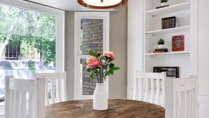 A round wooden table with white chairs, a white vase with flowers, and a brick wall in the background.