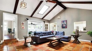 A spacious living area with a sectional sofa, a coffee table, and a dining table. There is a skylight above the sofa and a large window providing natural light.