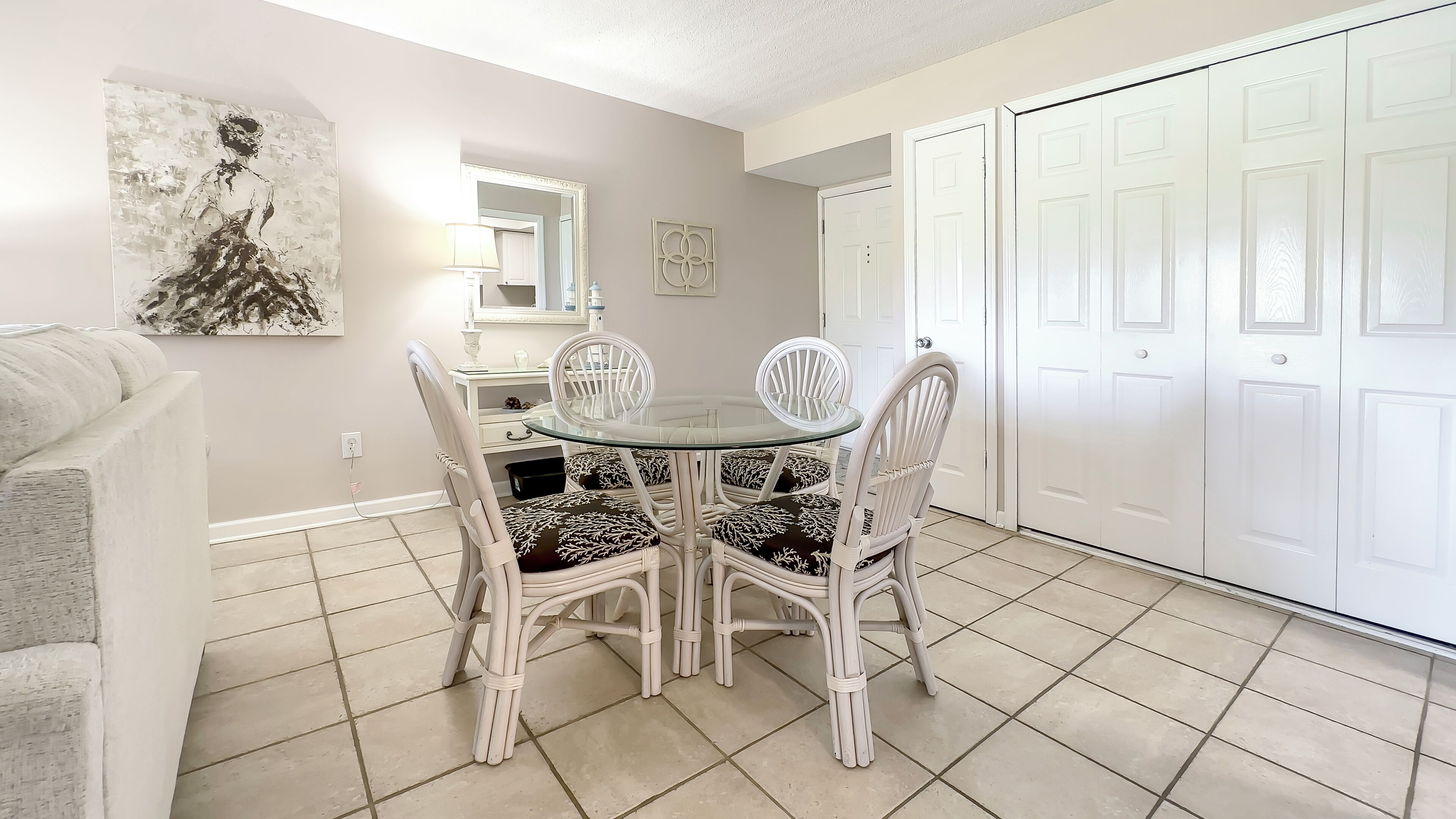 Un comedor con mesa de vidrio y sillas de mimbre blanco, una puerta blanca y piso de baldosas color beige.