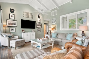 A living room with a flat-screen TV, a ceiling fan, and a large window with blinds.