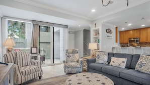 A modern living room with a grey sofa, striped armchair, and round ottoman, a kitchen with wooden cabinets, and a sliding glass door leading to a balcony. - 1261 Harbour Town Club at The Sea Pines Resort (Hilton Head Island)