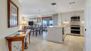 A modern kitchen with white cabinets, a built-in oven, and a microwave. There is a dining area with a table and chairs, and a view of the sea through the windows. - 1868 Beachside Tennis at The Sea Pines Resort (Hilton Head Island)