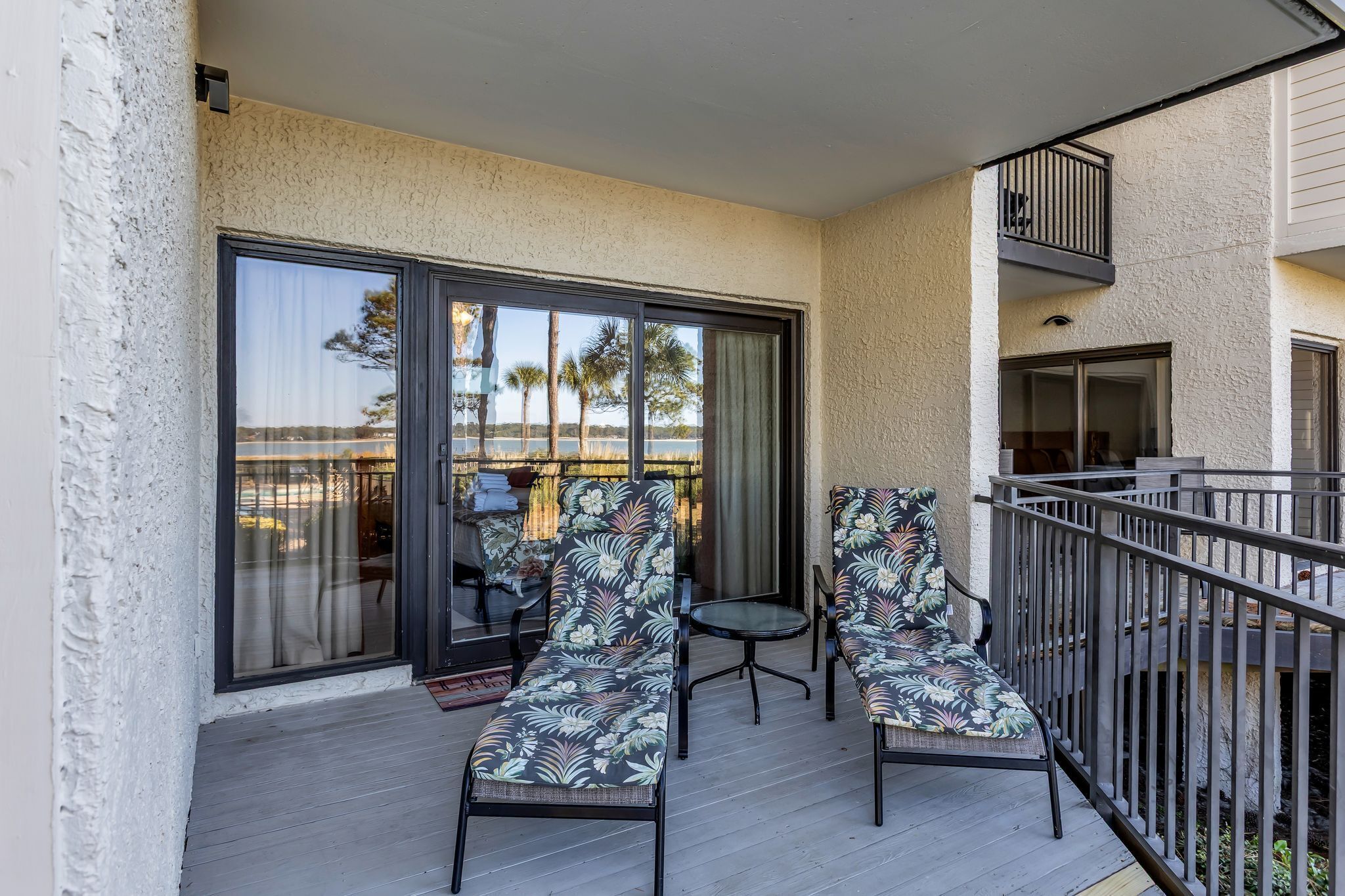 A balcony with two lounge chairs and a small table, a sliding glass door, and a view of palm trees.
