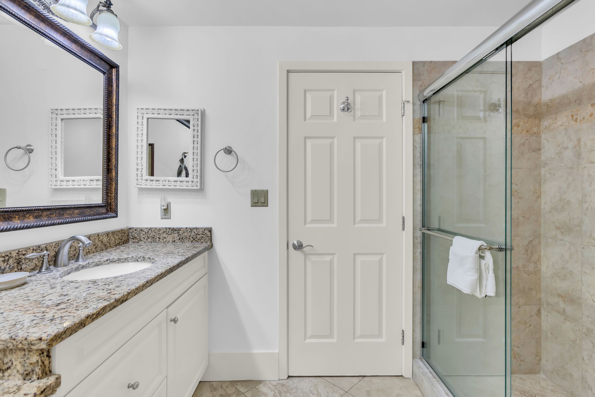 A bathroom with a granite countertop sink, two wall-mounted mirrors, a white door, and a glass shower enclosure.