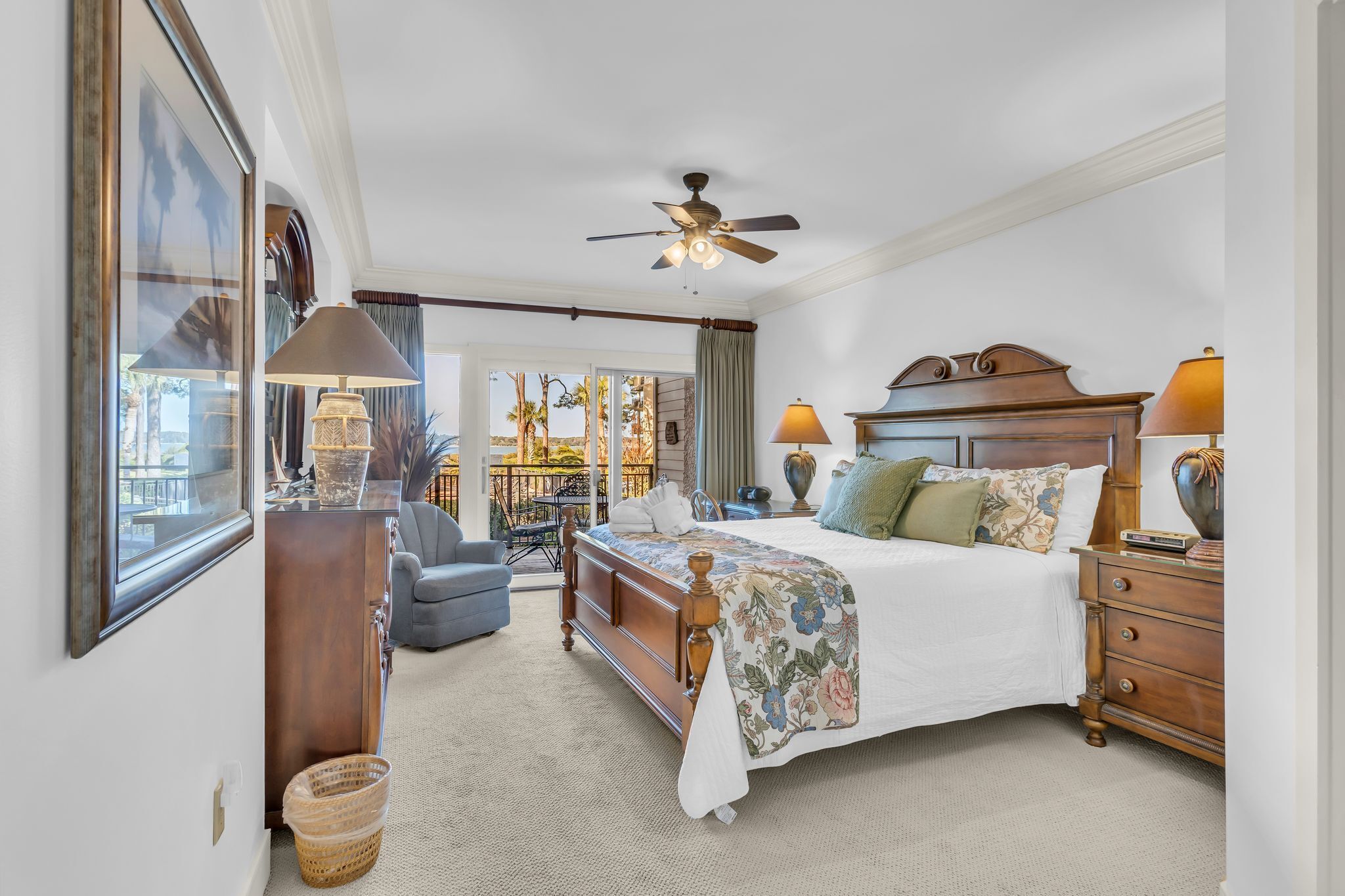 A spacious bedroom with a large bed, a wooden dresser, a desk with a lamp, and a view of a balcony with outdoor furniture.