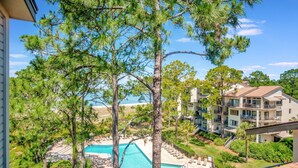 A pool area with lounge chairs, surrounded by pine trees and a view of a multi-story building with balconies.