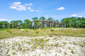 A multi-story hotel with a flat roof and balconies, surrounded by a natural landscape with trees and sandy terrain.