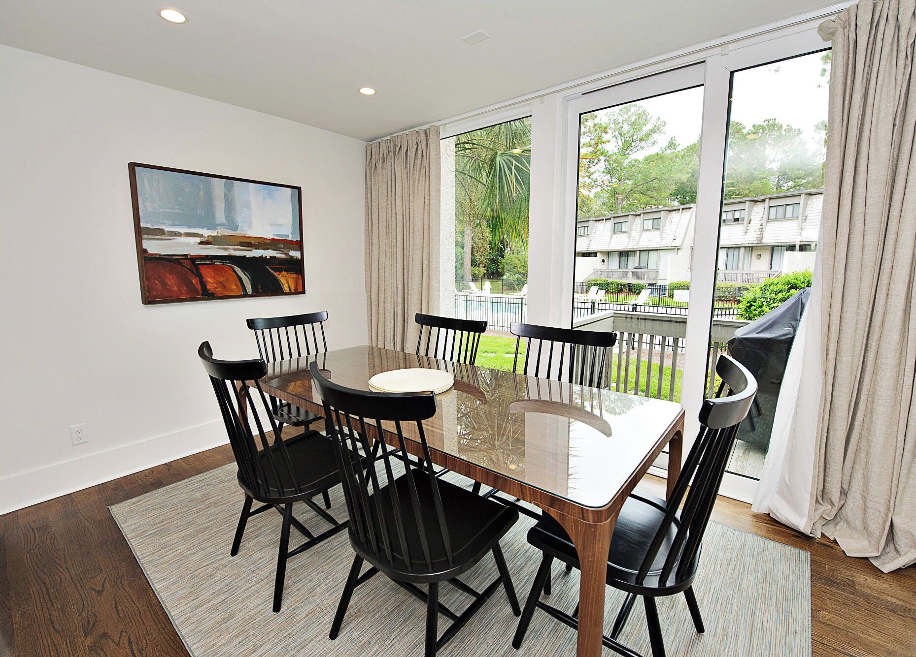 Uma área de refeições com mesa de vidro e cadeiras pretas, um quadro na parede e vista para uma área residencial através de grandes janelas.