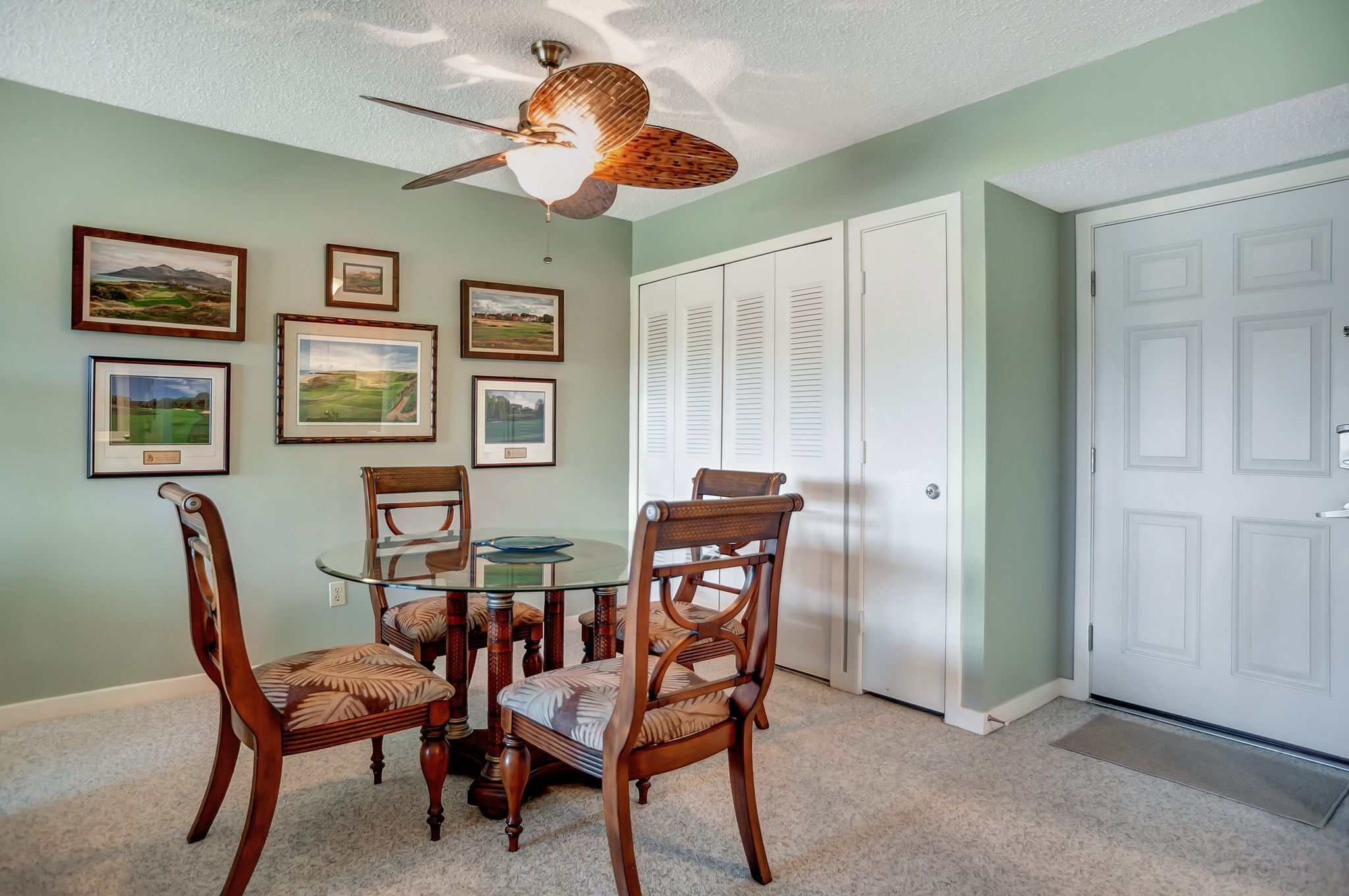 A dining area with a glass table and wooden chairs, a ceiling fan, and framed pictures on the wall.
