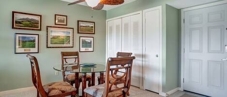 A dining area with a glass table and wooden chairs, a ceiling fan, and framed pictures on the wall.