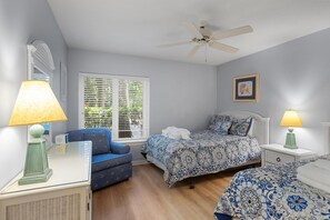 Ein Schlafzimmer mit einem Bett, einem Sessel, einem Nachttisch, einer Lampe, einem Deckenventilator und einem Fenster mit Jalousien.