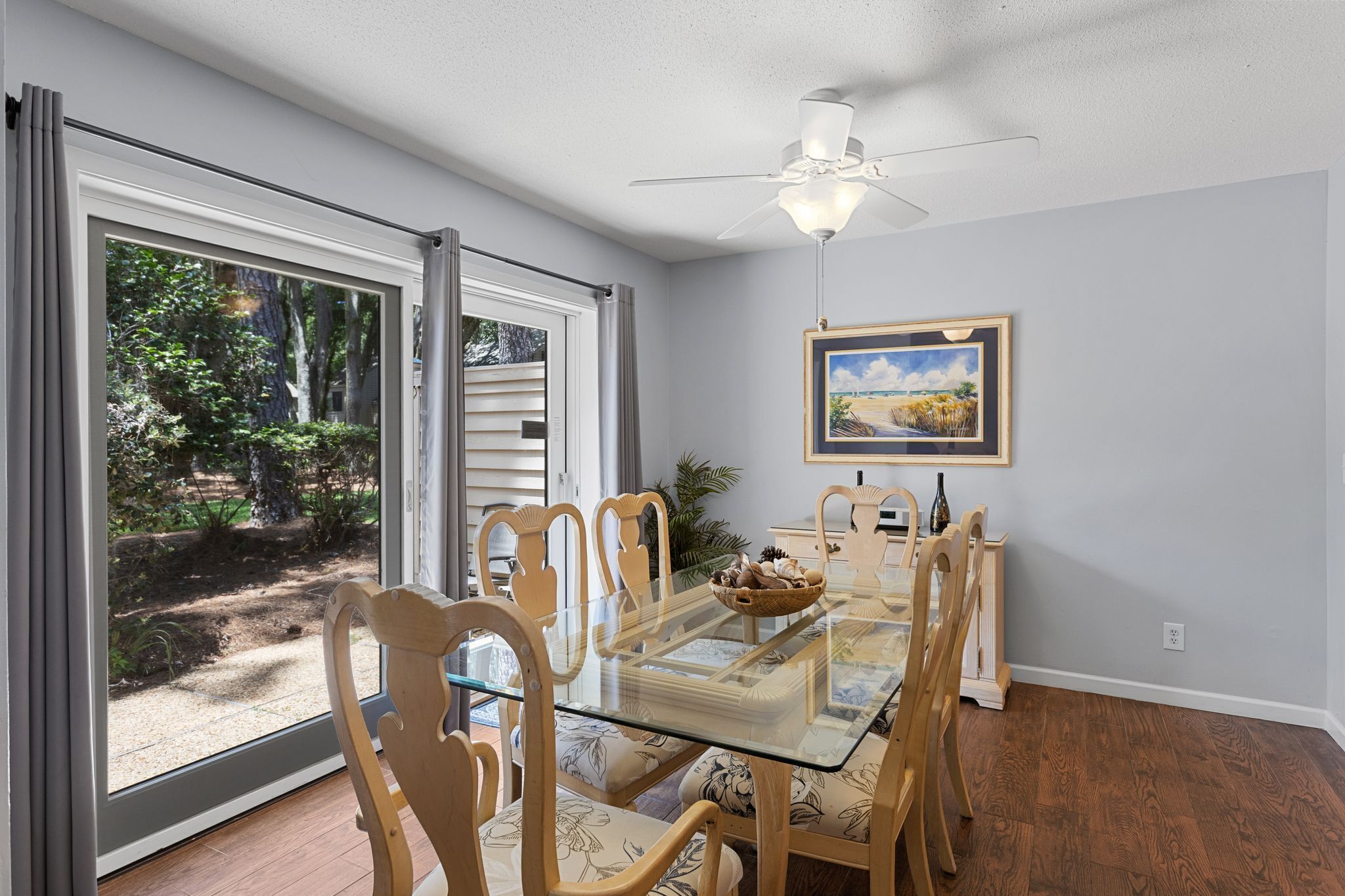 A dining area with a glass table, wooden chairs, a ceiling fan, and a painting on the wall.
