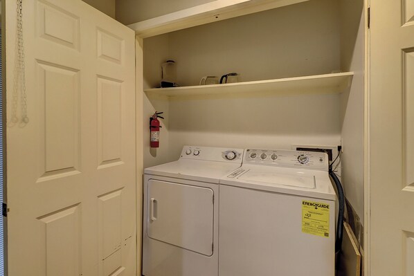 A compact laundry room with a front-loading washer and dryer, a fire extinguisher, and a shelf above the dryer.