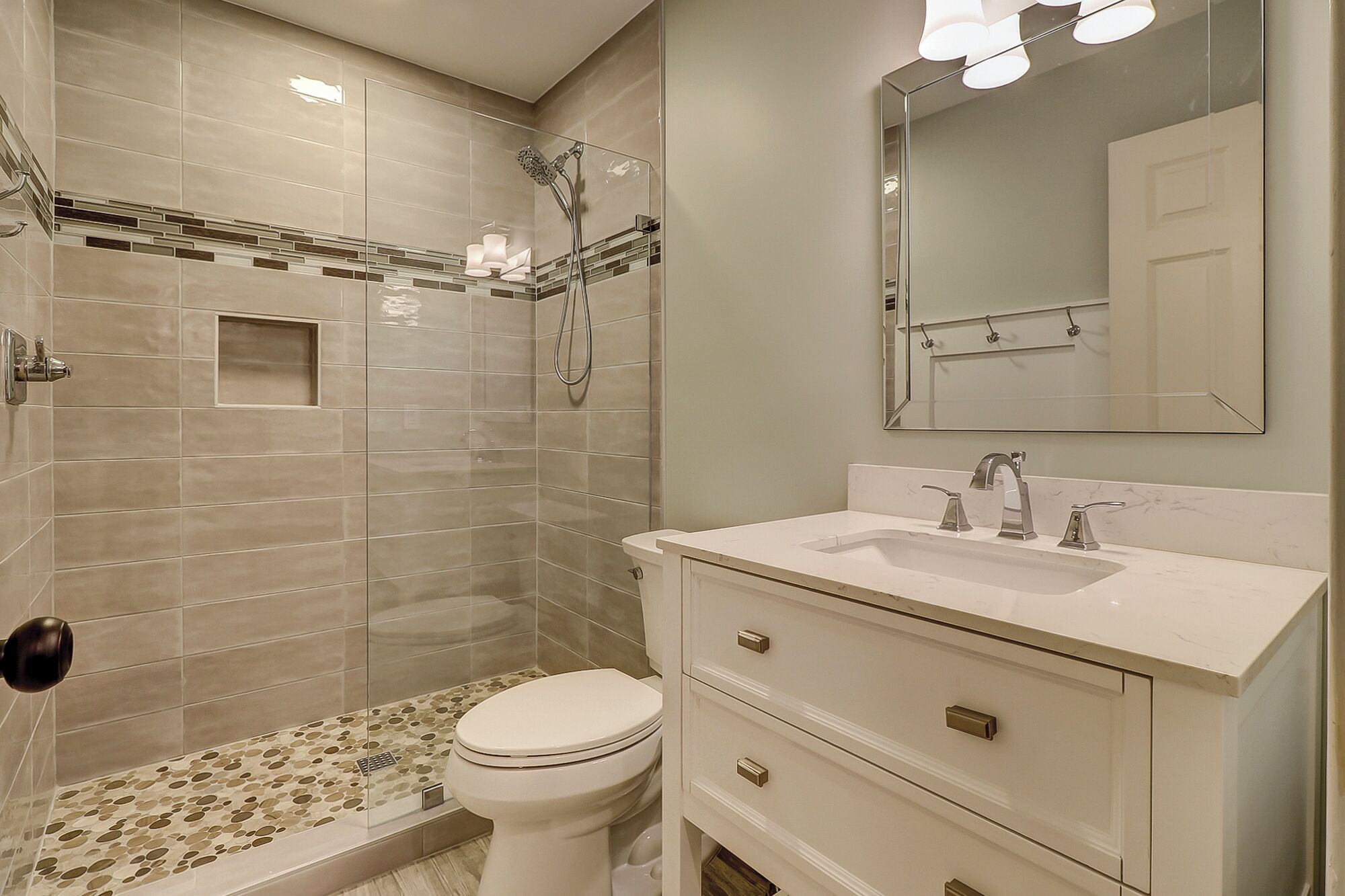 Un baño moderno con cabina de ducha de vidrio, inodoro blanco, tocador con lavamanos y espejo, y suelo de baldosas.